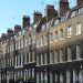 Image showing a row of Georgian-style brown brick terraced houses with uniform chimneys and white-framed windows, located on a street under a clear blue sky. People are walking on the sidewalk in front of the buildings. | TitaniumInvest