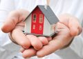 A person holds a small model house in their cupped hands. The house is white with red accents and a grey roof. The background is blurred, focusing on the house and hands. | TitaniumInvest
