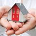 A person holds a small model house in their cupped hands. The house is white with red accents and a grey roof. The background is blurred, focusing on the house and hands. | TitaniumInvest