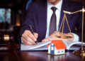 A person in a suit, likely a real estate attorney, is writing in a notebook at a desk. In the foreground, there is a small model house, a gavel, and a pair of scales. The scene suggests a legal or real estate context. The background is blurred. | TitaniumInvest