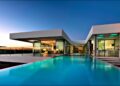 Modern house with large glass windows and a sleek design, overlooking an infinity pool under a twilight sky. Inside, the contemporary decor is visible through the windows, featuring a spacious living area and illuminated kitchen with circular ceiling lights. Perfect luxury real estate in Portugal. | TitaniumInvest