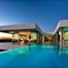 Modern house with large glass windows and a sleek design, overlooking an infinity pool under a twilight sky. Inside, the contemporary decor is visible through the windows, featuring a spacious living area and illuminated kitchen with circular ceiling lights. Perfect luxury real estate in Portugal. | TitaniumInvest