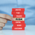 A person wearing a blue shirt is pushing a block labeled "RISK" in the middle of a stack of red blocks with the words "ANALYSIS," "PLAN," "EVALUATE," "CONTROL," "REVIEW," and "ASSESSMENT" written on each block. | TitaniumInvest