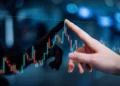 A close-up of a hand pointing at a digital screen displaying a candlestick chart with red and green bars, indicating stock or financial data. The background is blurred with blue hues and light spots, suggesting a busy, high-tech investing environment. | TitaniumInvest