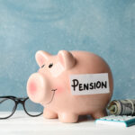 A piggy bank labeled "Pension" sits on a wooden surface, embodying financial wellness. Nearby, a pair of glasses rests beside a small calculator and a rolled-up bundle of cash, set against a textured blue background. | TitaniumInvest