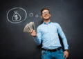 A man in a light blue shirt and jeans stands smiling in front of a blackboard. As a TitaniumInvest.com Money Expert, he holds a fan of dollar bills in one hand while above his head is a chalk drawing of a money bag marked with a dollar sign. | TitaniumInvest