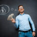 A man in a light blue shirt and jeans stands smiling in front of a blackboard. As a TitaniumInvest.com Money Expert, he holds a fan of dollar bills in one hand while above his head is a chalk drawing of a money bag marked with a dollar sign. | TitaniumInvest