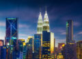 Kuala Lumpur city skyline at dusk, featuring brightly lit skyscrapers including the iconic Petronas Twin Towers and Kuala Lumpur Tower against a deep blue sky with streaks of clouds. | TitaniumInvest