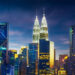 Kuala Lumpur city skyline at dusk, featuring brightly lit skyscrapers including the iconic Petronas Twin Towers and Kuala Lumpur Tower against a deep blue sky with streaks of clouds. | TitaniumInvest