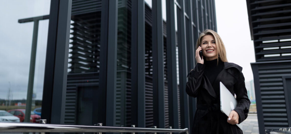 A woman in a black coat stands outside modern office buildings, smiling while talking on her phone and holding a closed laptop under her arm. For more information, contact TitaniumInvest.com. | TitaniumInvest
