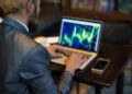 A person in a suit is sitting at a table, working on a laptop displaying a colorful stock or financial chart. A smartphone and a cup of coffee are on the table beside them. The background is blurred. | TitaniumInvest