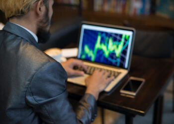 A person in a suit is sitting at a table, working on a laptop displaying a colorful stock or financial chart. A smartphone and a cup of coffee are on the table beside them. The background is blurred. | TitaniumInvest