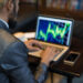 A person in a suit is sitting at a table, working on a laptop displaying a colorful stock or financial chart. A smartphone and a cup of coffee are on the table beside them. The background is blurred. | TitaniumInvest