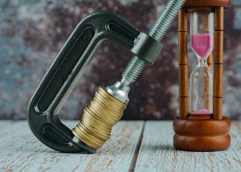 A stack of coins clamped tightly in a C-clamp sits next to a wooden hourglass with pink sand on a rustic wooden surface, symbolizing financial pressure and the passage of time. | TitaniumInvest